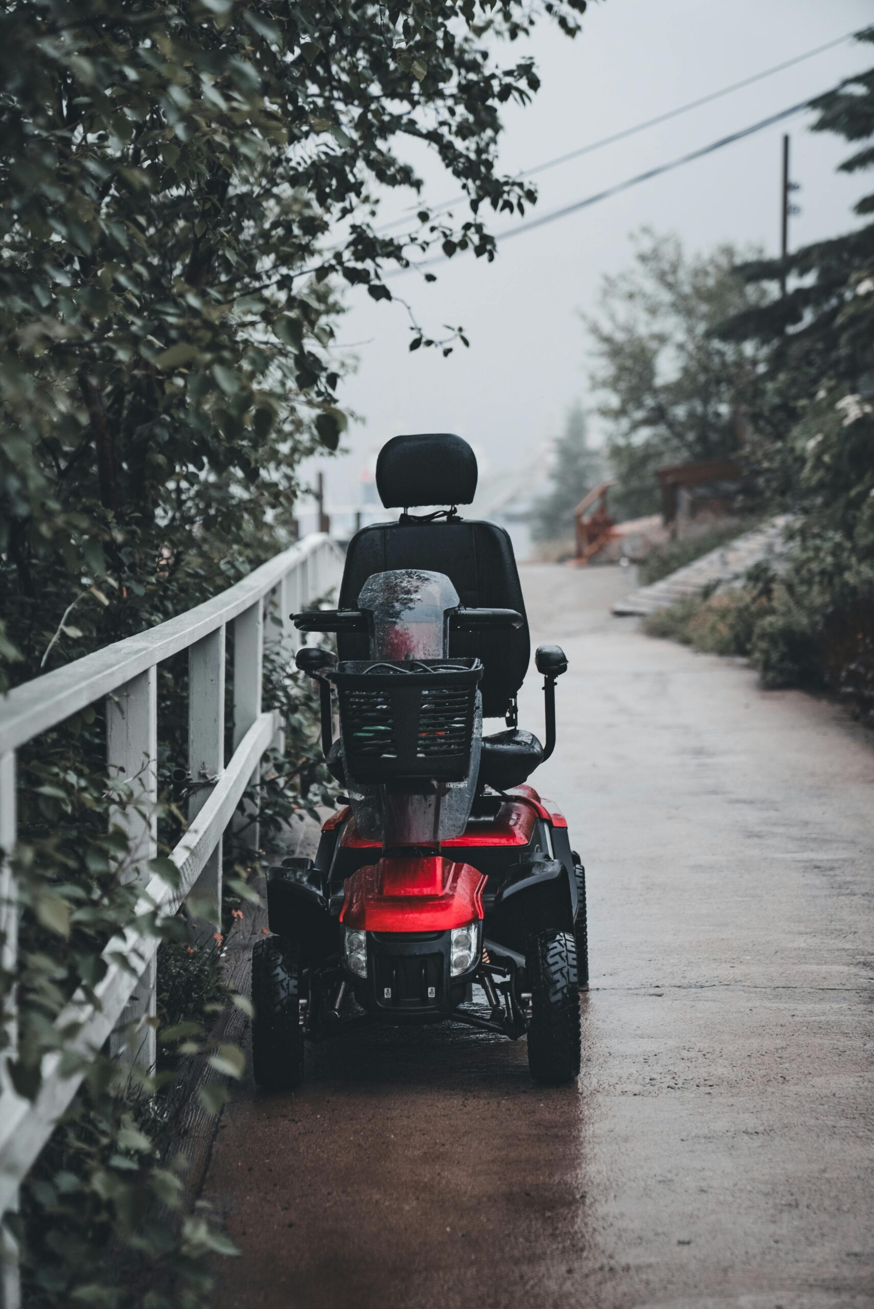 A red mobility scooter parked on a rainy village street path lined with trees and railings.